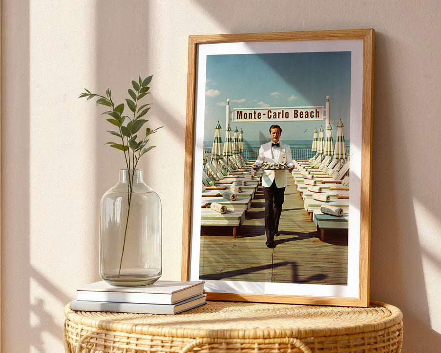 Framed photograph of a man on a beach with 'Monte-Carlo Beach' sign, placed on a table with a vase and books.