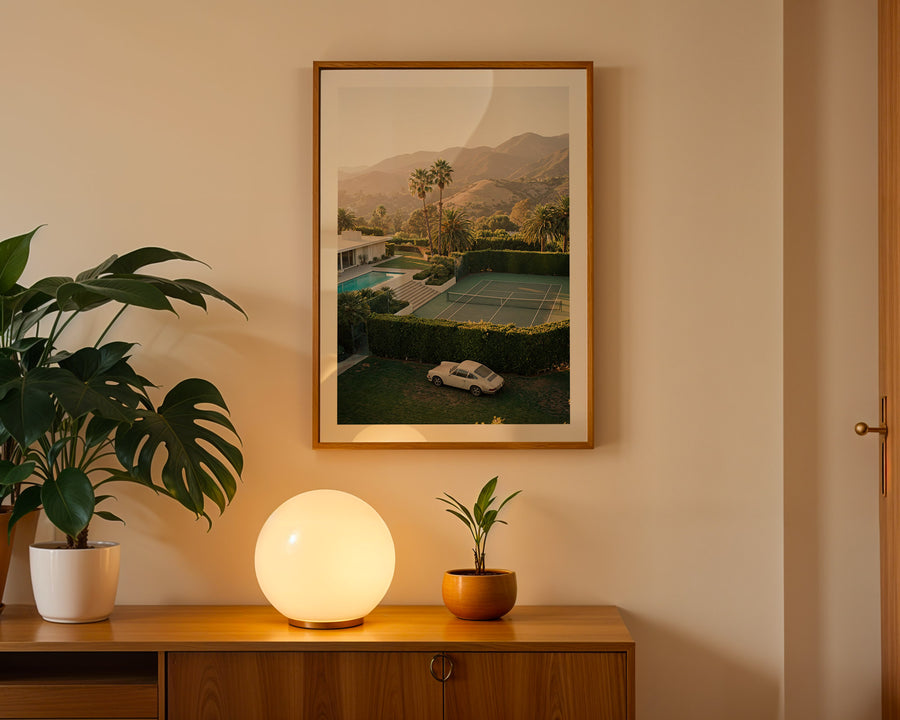 Framed artwork of a tennis court on a wall with a plant and lamp on a wooden surface below.