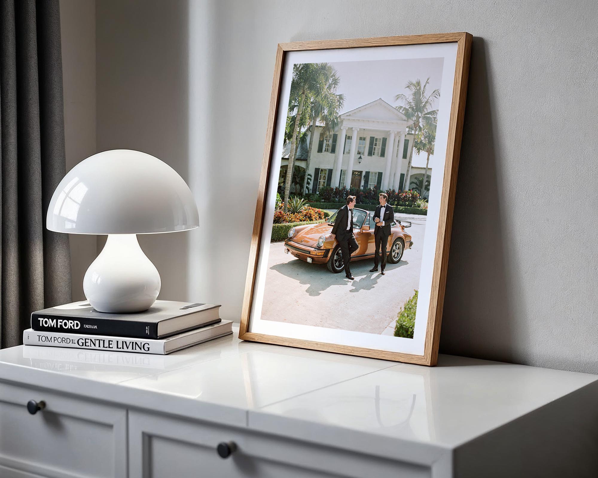 Framed photograph of two people by a vintage car on a white surface with a lamp and books.