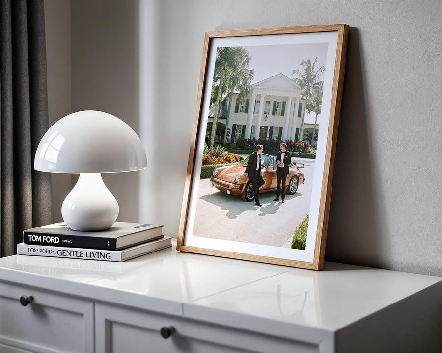 Framed photograph of two people by a vintage car on a white surface with a lamp and books.
