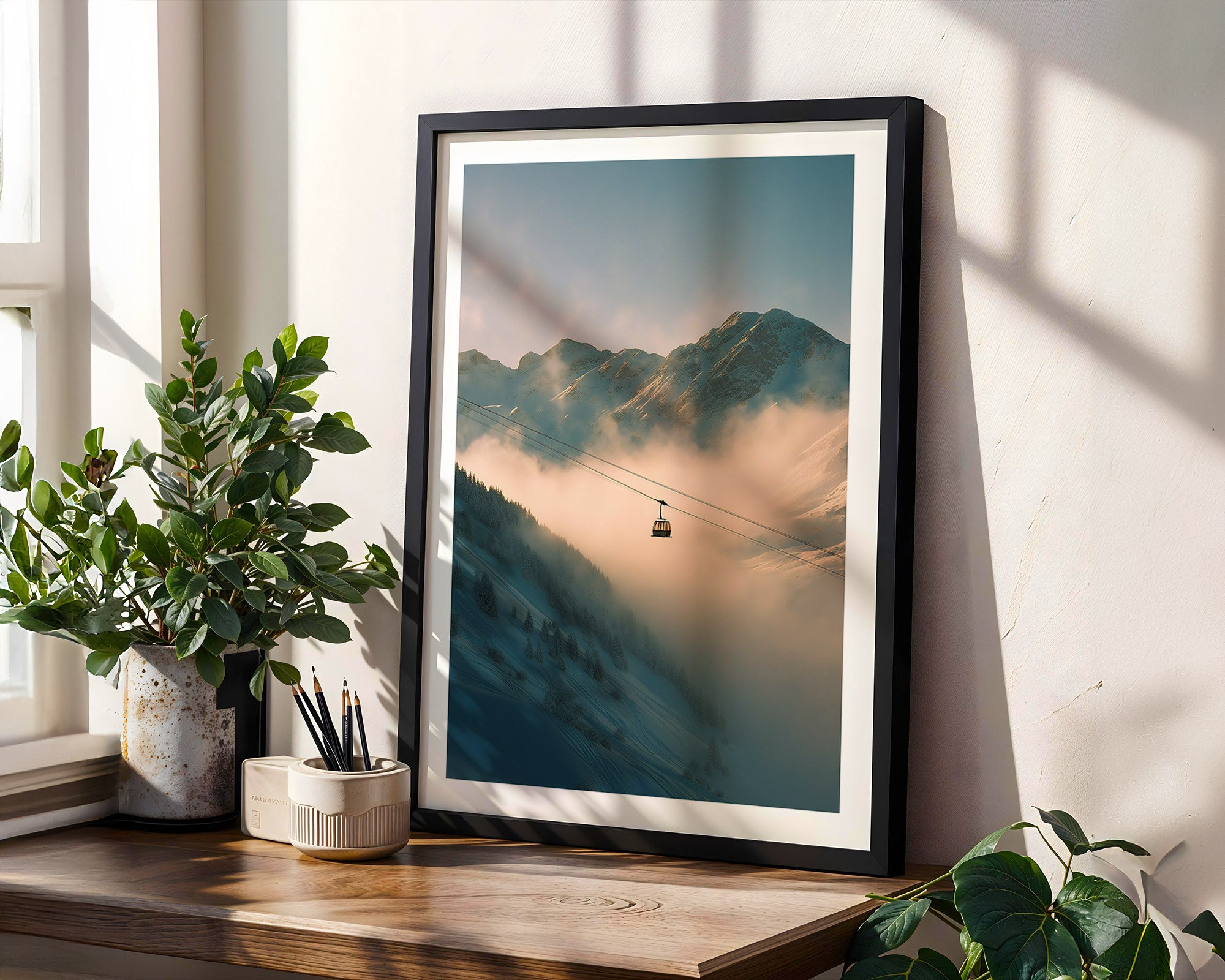 Framed mountain landscape print on a wall with plants and a window in the background