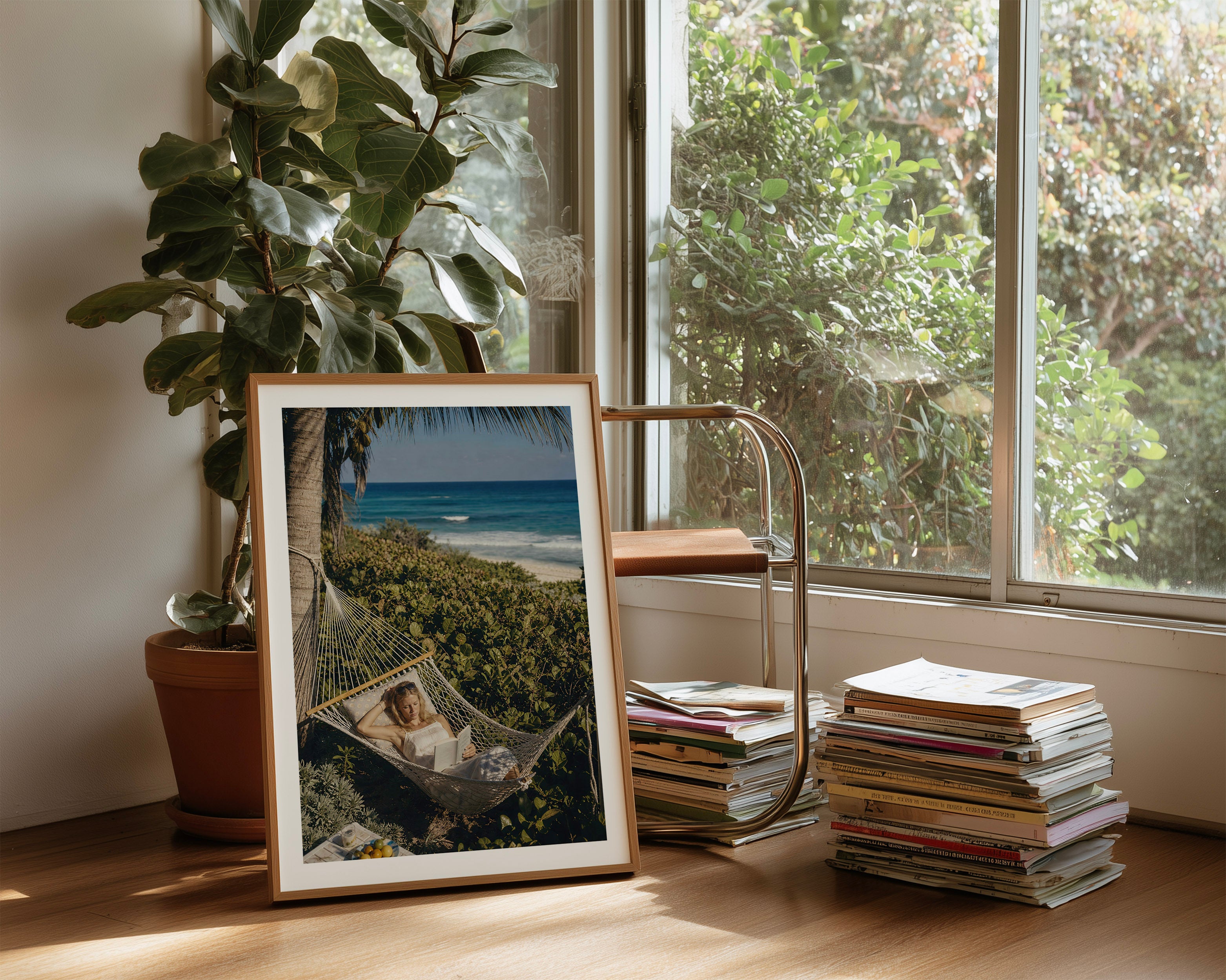 A framed vertical art print leaning on a wooden floor in a sunlit room. The print shows a woman relaxing in a hammock outdoors near a beach and ocean.