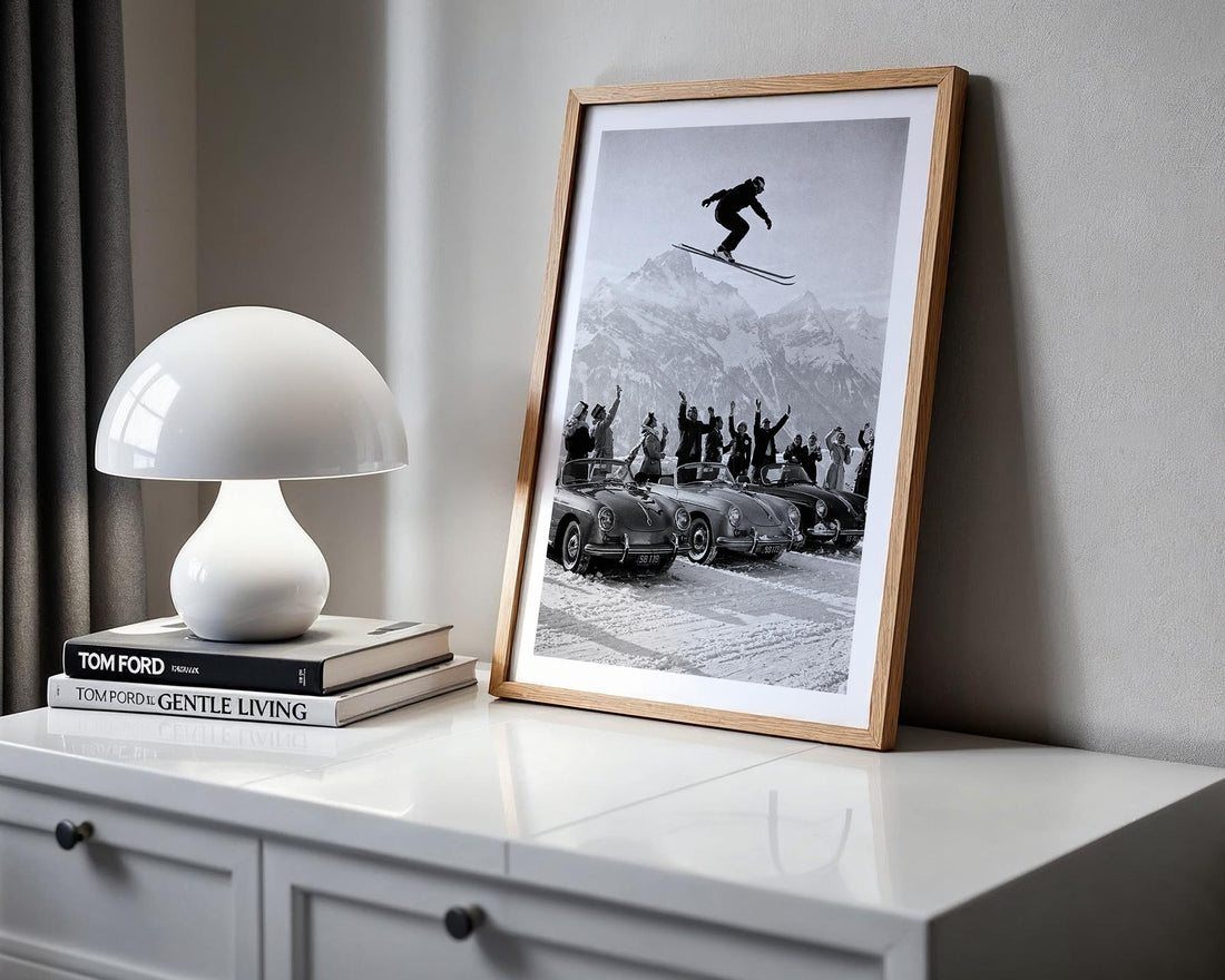 Framed black and white photo of a skier above a military vehicle on a shelf with a lamp and books.