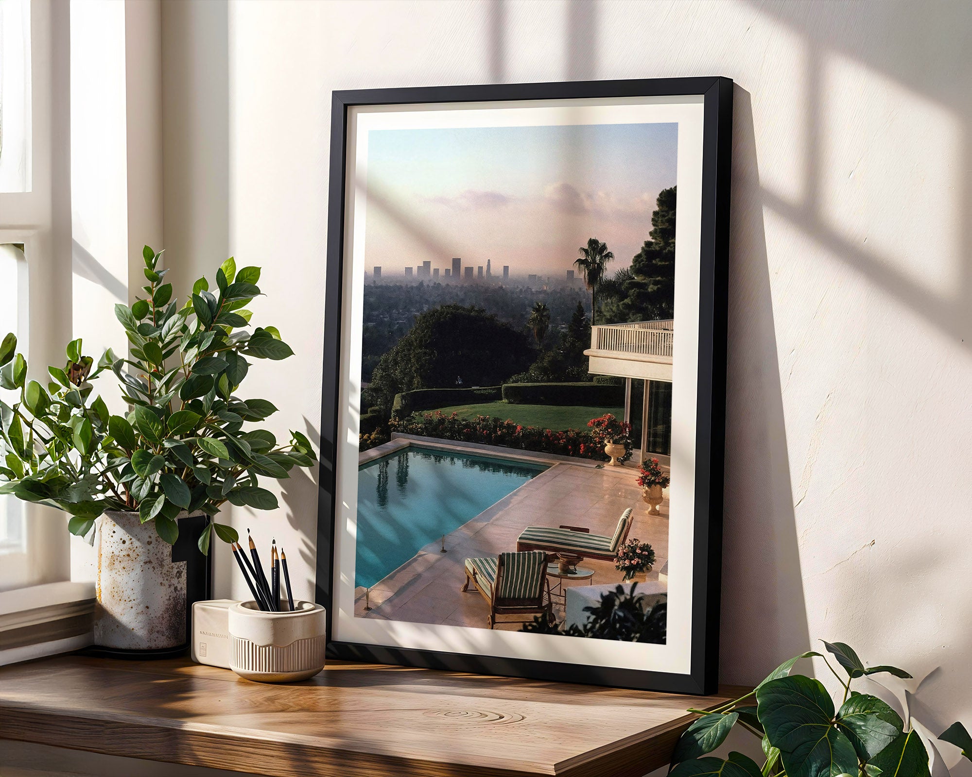 Framed artwork of a poolside scene in Los Angeles on a wall with plants and decor below.