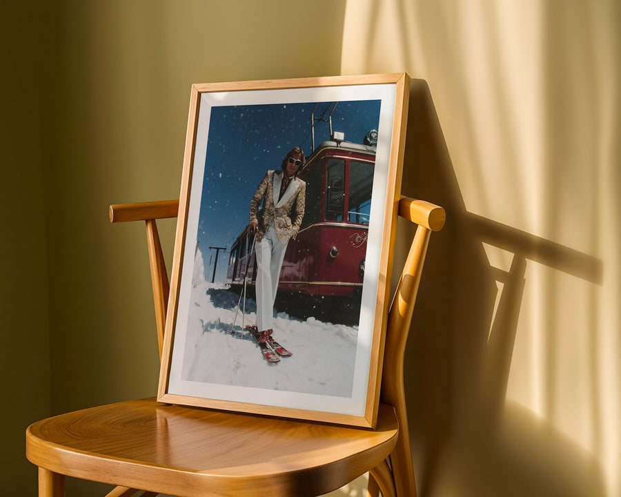 Framed photograph of a person skiing next to a red vehicle on a wooden chair.