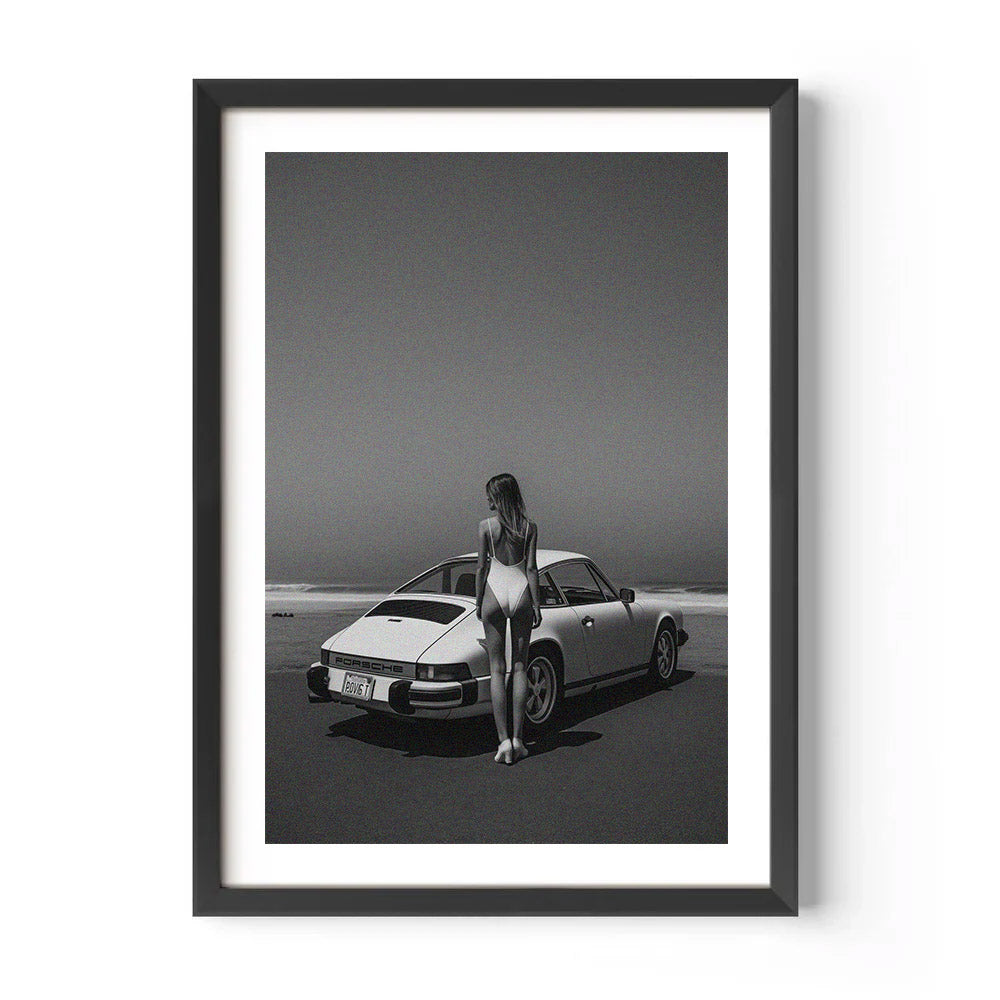 Framed black and white photo of a person standing next to a vintage car on a beach.