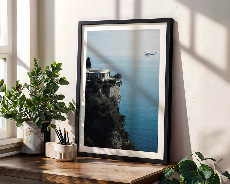 Framed artwork of a coastal scene with a helicopter over water, placed on a wooden surface with plants.