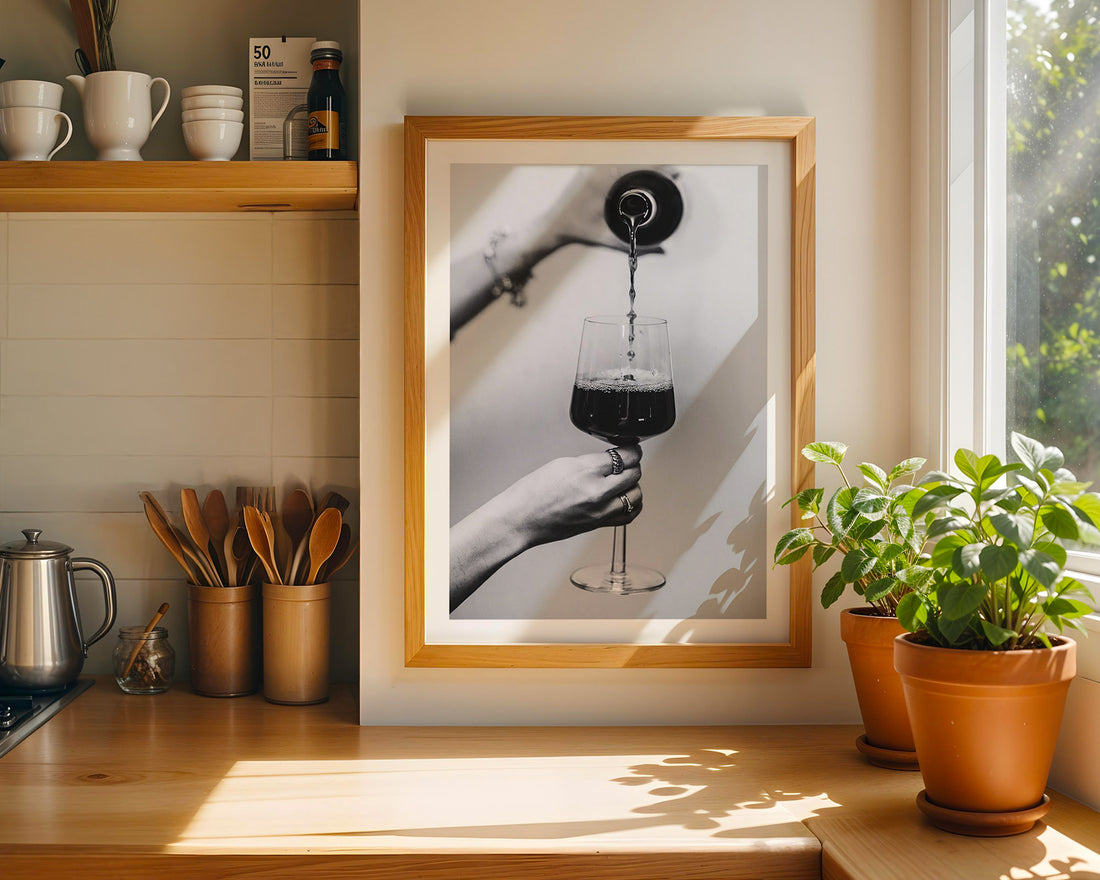 Framed artwork of a hand pouring liquid into a glass on a kitchen counter with plants and coffee cups.