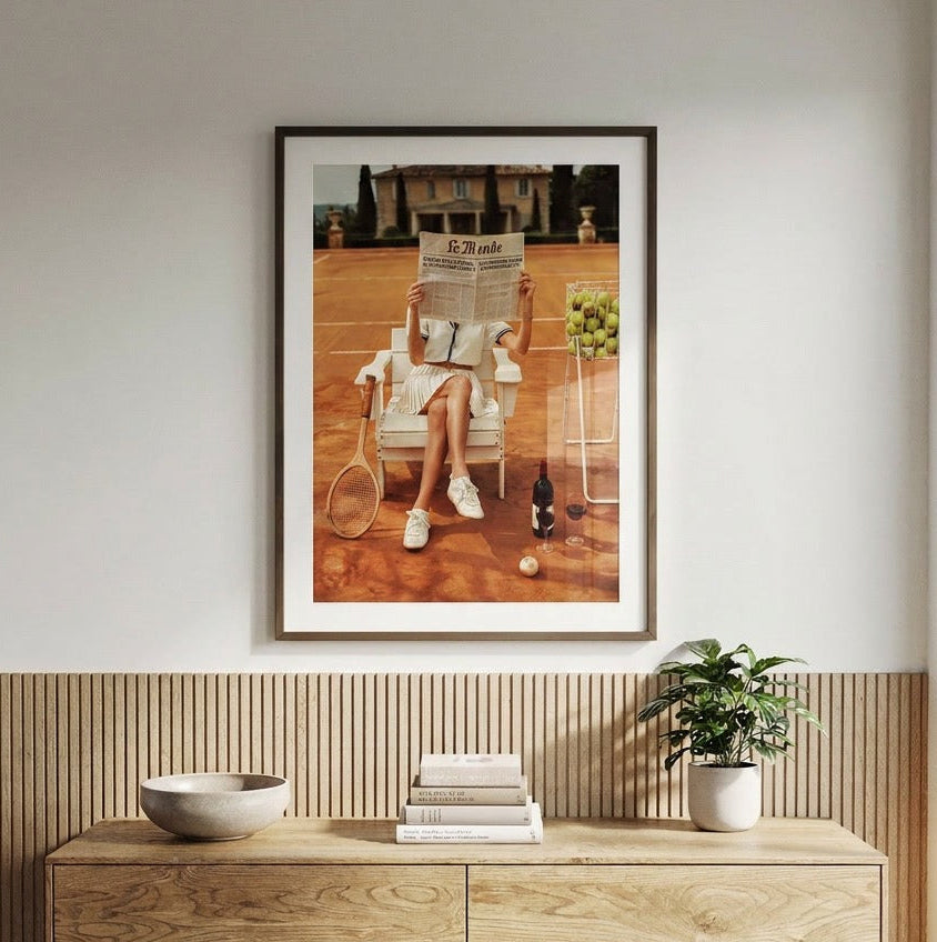 A framed art print depicting a woman sitting on a tennis court reading a newspaper, displayed on a light gray wall above a wooden console table with a bowl, books, and a potted plant.
