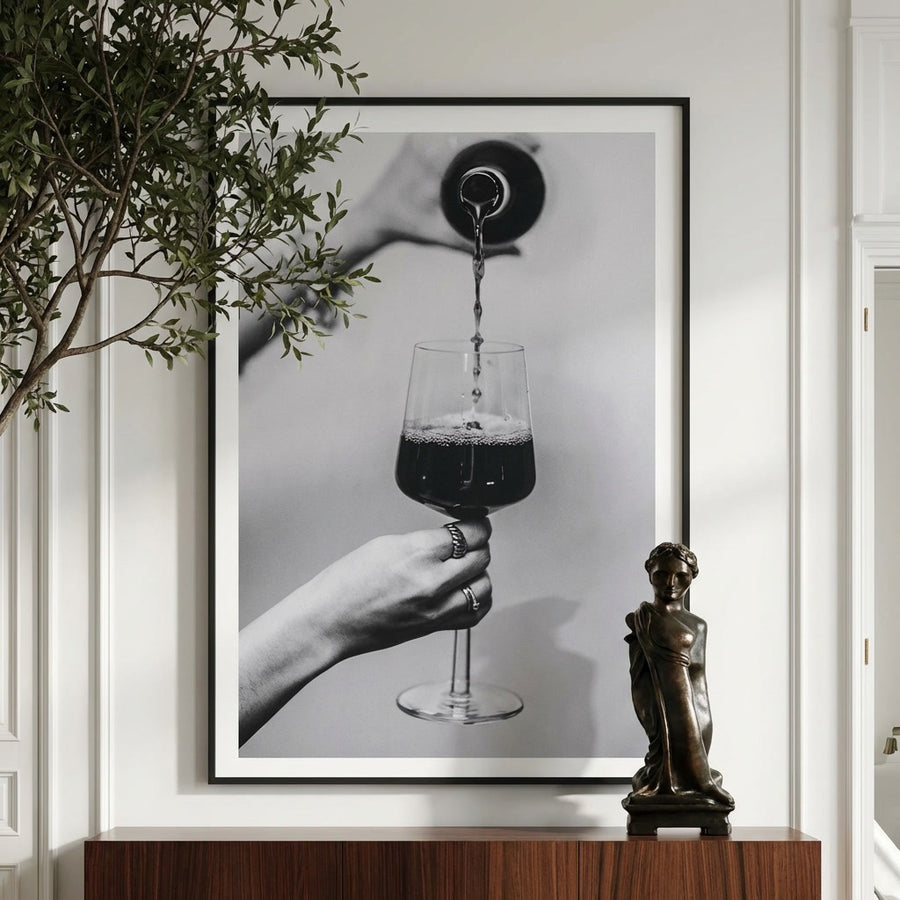 Framed artwork of a hand holding a wine glass with red wine, placed on a shelf with a plant and statue.