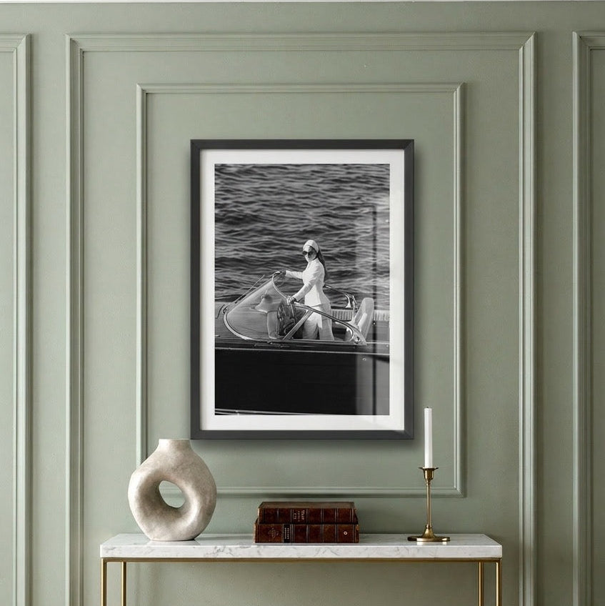 A framed black and white art print of a person rowing on water, displayed on a green paneled wall above a console table with a white vase, books, and a candle.
