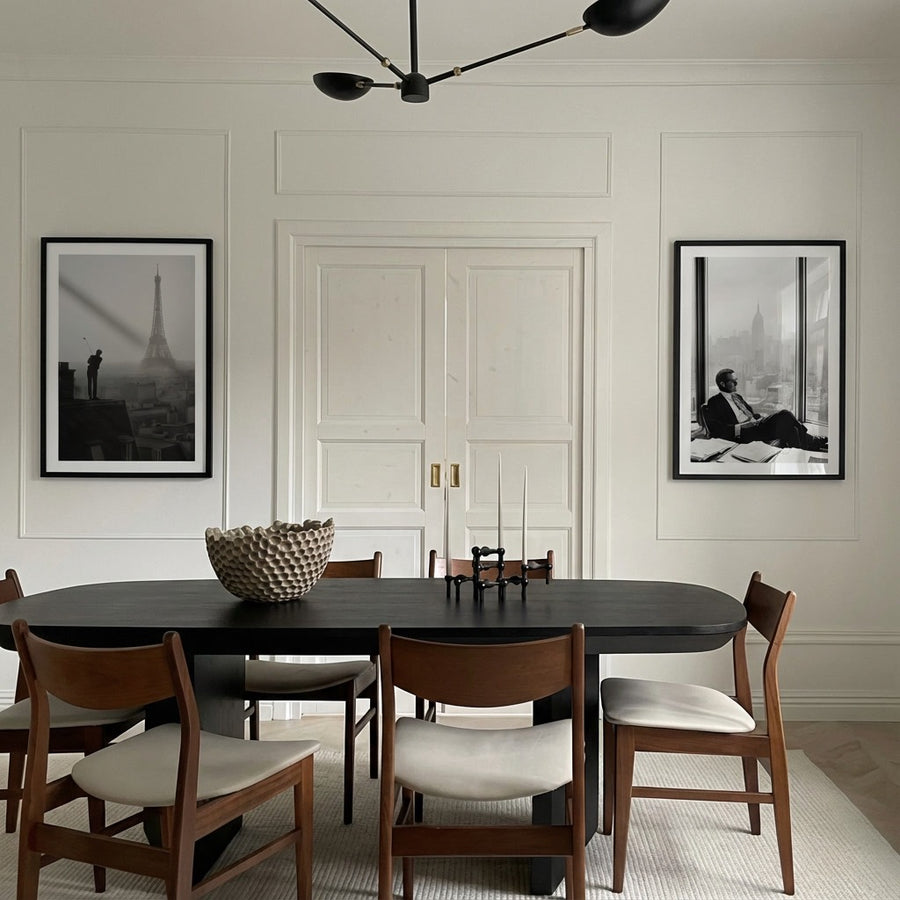 Dining room with a black oval table and wooden chairs with white cushions, framed pictures on the wall.