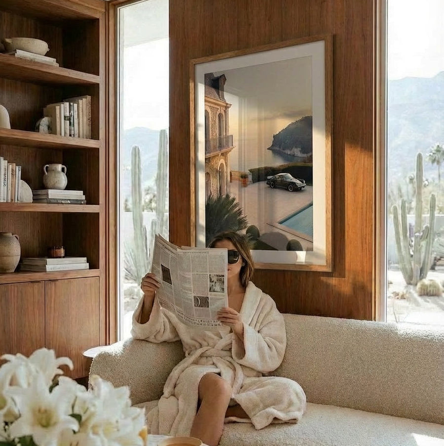 Person in a robe reading a newspaper in a cozy living room with cacti outside the window.