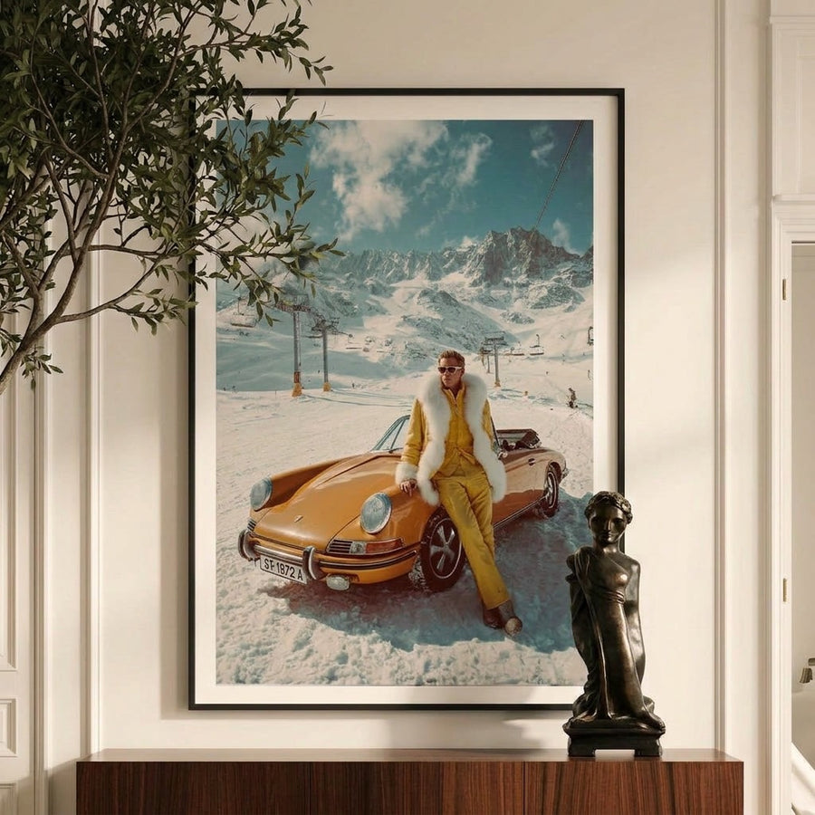 Framed artwork of a person in a yellow outfit sitting on a vintage car in a snowy landscape, displayed in a room with a plant and sculpture.