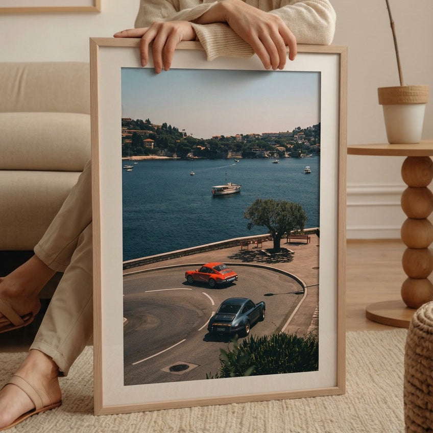 Person holding a framed photograph of a coastal scene with cars and a boat.