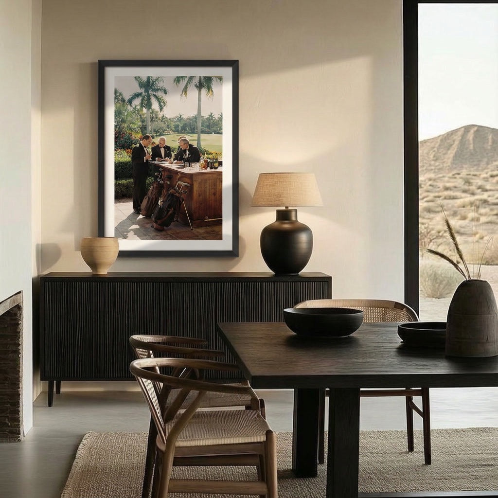A framed art print depicting a vintage scene of people at a table in Palm Springs, displayed on a dark wood sideboard in a modern dining room setting.