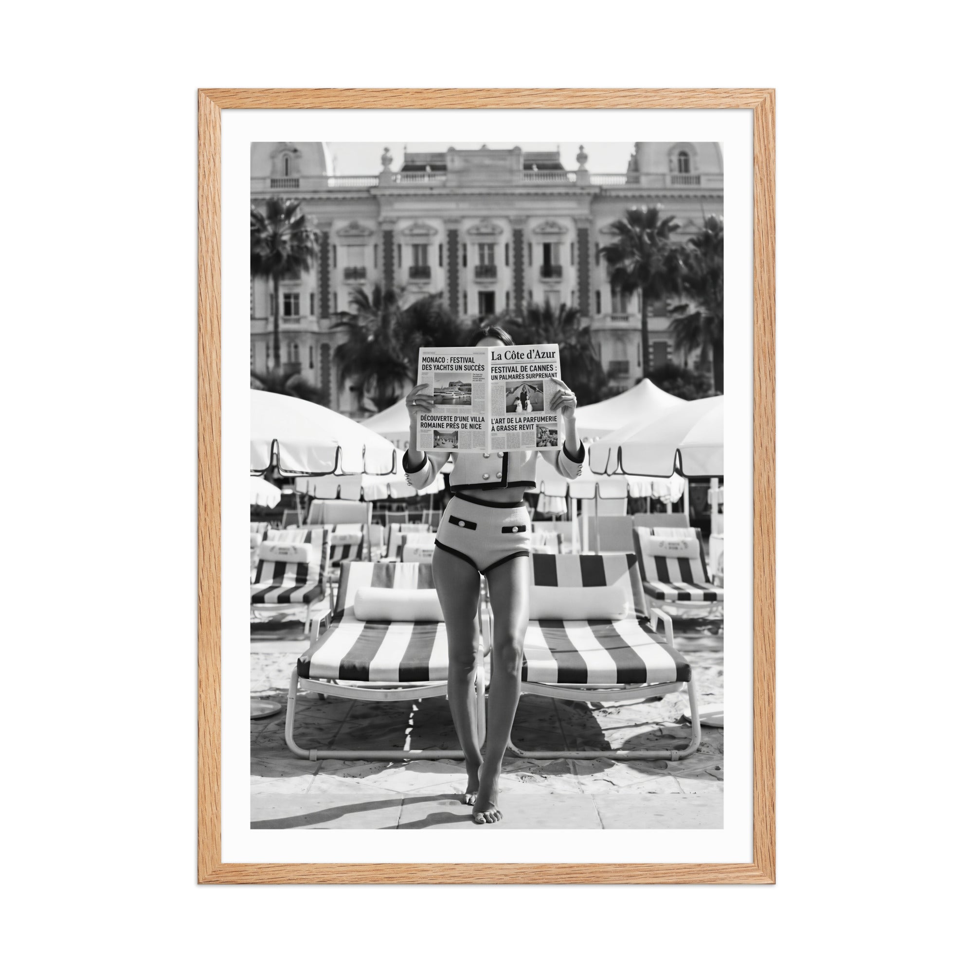 Framed black and white photo of a woman in a bikini reading a newspaper on a striped chair.