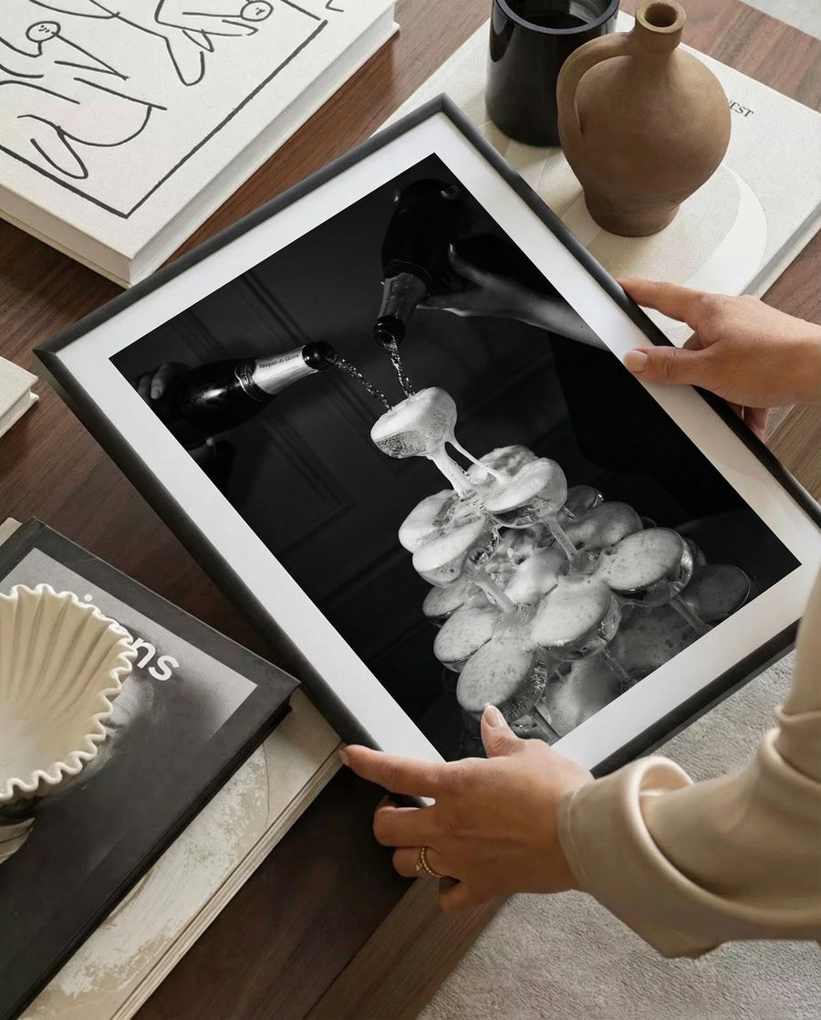 Person holding a framed black and white photograph of a wine bottle being poured into glasses.